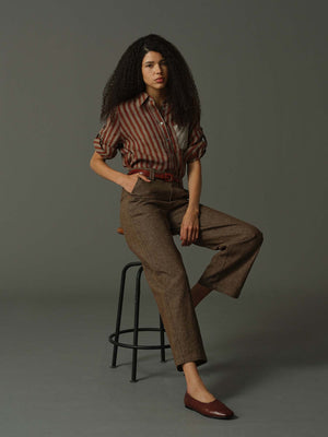 Woman in a striped shirt and brown pants sitting on a stool against a gray background