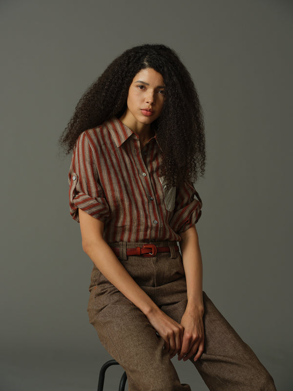 Woman sitting on a stool wearing a striped shirt and brown pants against a gray background