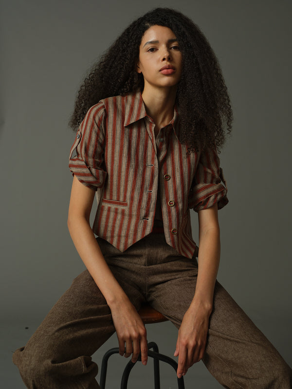 Woman wearing a striped shirt and brown pants sitting on a stool against a gray background