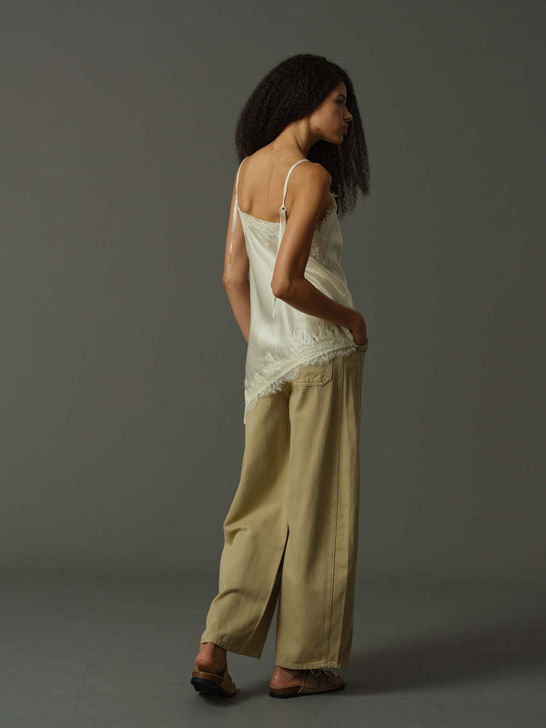 Woman wearing a light-colored lace top and beige pants against a gray background