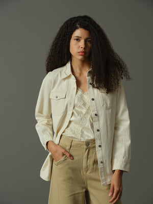 Woman wearing a light-colored jacket over a lace top and beige pants on a gray background