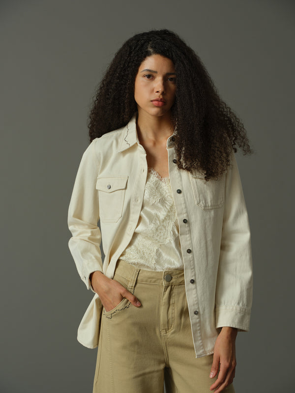 Woman wearing a light-colored jacket over a lace top and beige pants on a gray background