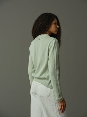 Woman wearing a light green sweater and white pants against a gray background