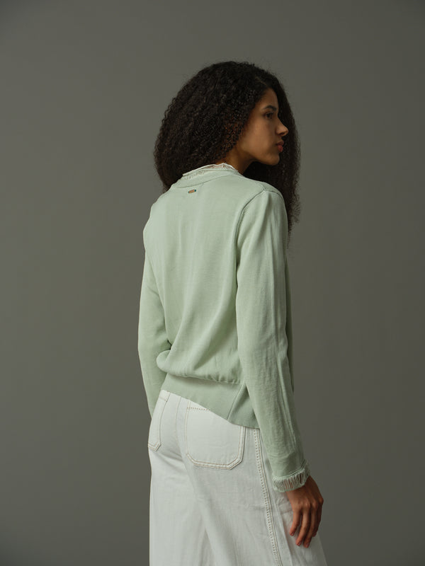 Woman wearing a light green sweater and white pants against a gray background