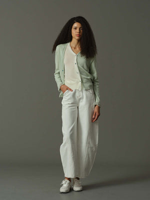 Woman wearing a light green cardigan, white shirt, and white pants against a gray background