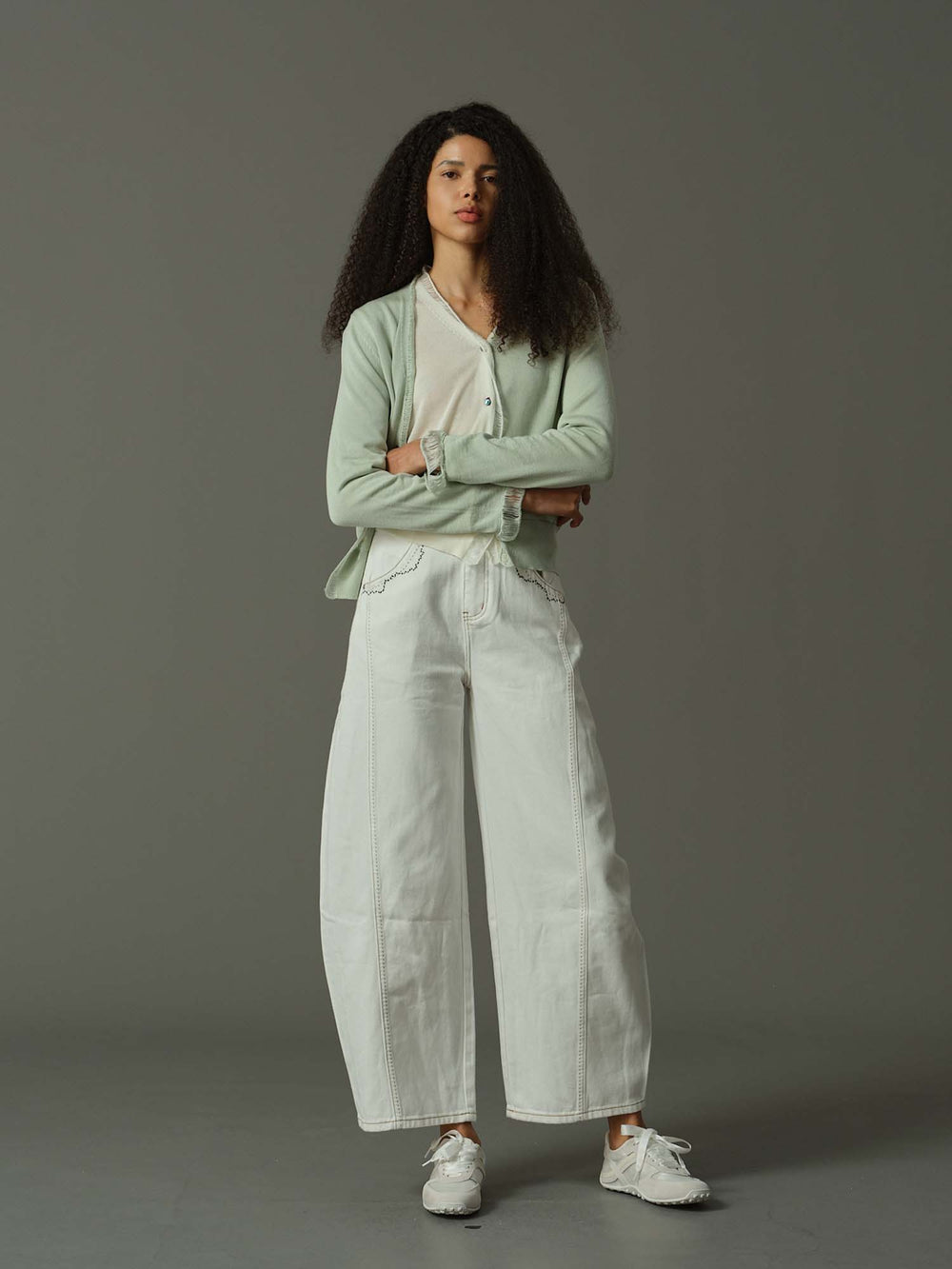 Woman wearing a light green cardigan and white pants against a gray background