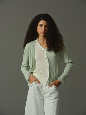 Woman wearing a light green cardigan over a white shirt and light blue jeans against a gray background