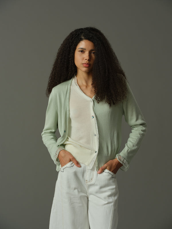 Woman wearing a light green cardigan over a white shirt and light blue jeans against a gray background