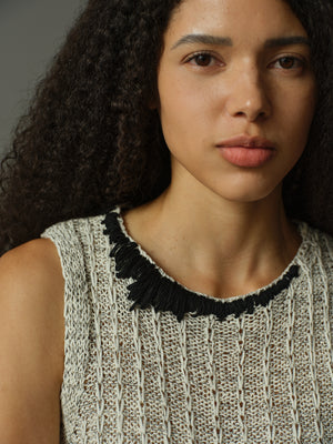 Woman wearing a sleeveless knitted top with a black fringed neckline against a neutral background