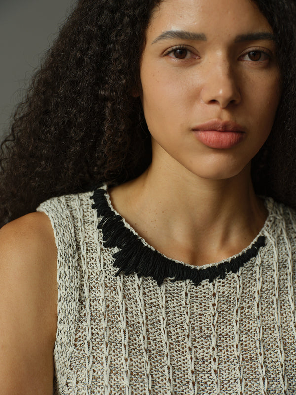 Woman wearing a sleeveless knitted top with a black fringed neckline against a neutral background