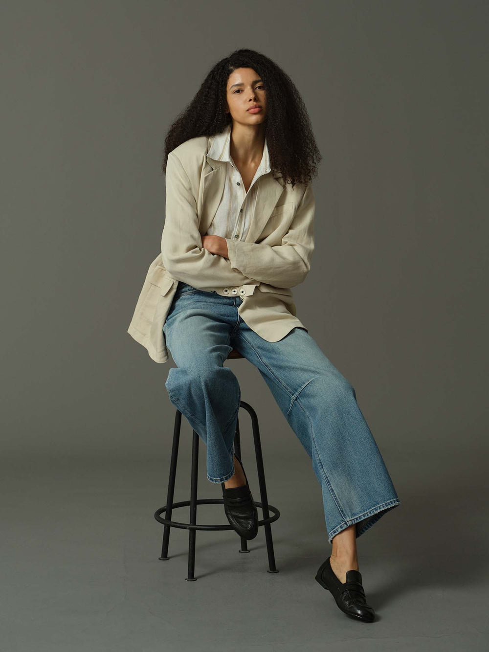 Woman sitting on a stool wearing a beige jacket, white shirt, blue jeans, and black shoes against a gray background.