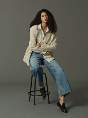 Woman sitting on a stool wearing a beige jacket, white shirt, blue jeans, and black shoes against a gray background.
