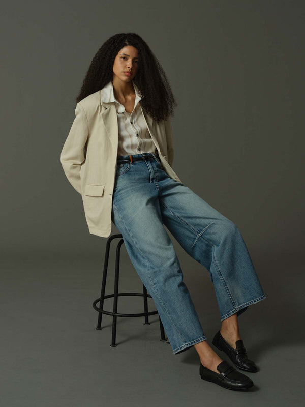 Woman sitting on a stool wearing a beige blazer, white shirt, blue jeans, and black shoes against a gray background.