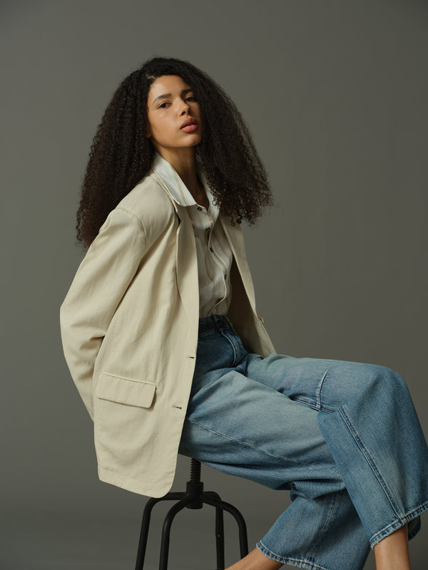 Woman wearing a beige blazer and blue jeans sitting on a stool against a gray background