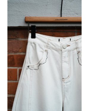 White denim jeans with lace details hanging on a wooden rod against a brick wall.