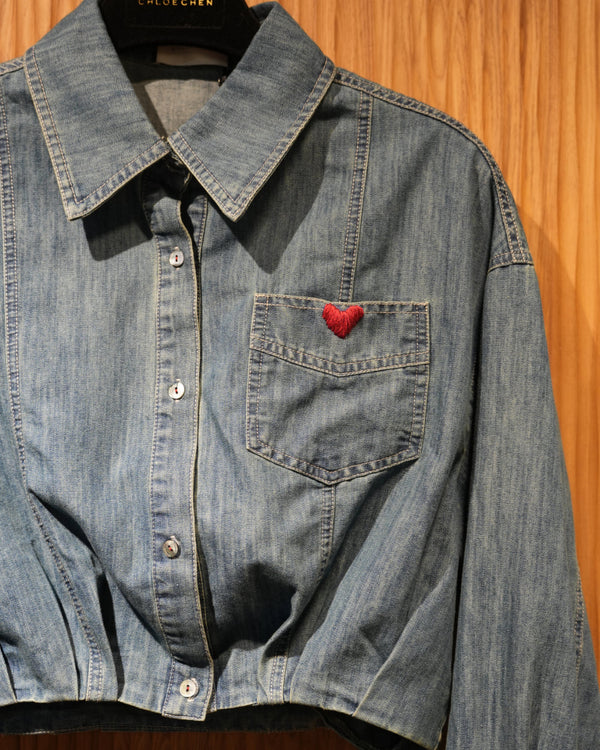 cropped denim jacket with heart embroidery on chest pocket, hanging on a brown textured wall