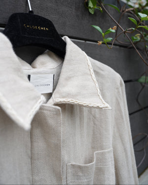 close up of a linen jacket with braided collar details on a hanger with a visible tag, against a dark surface with greenery.