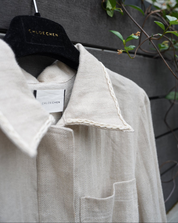 close up of a linen jacket with braided collar details on a hanger with a visible tag, against a dark surface with greenery.