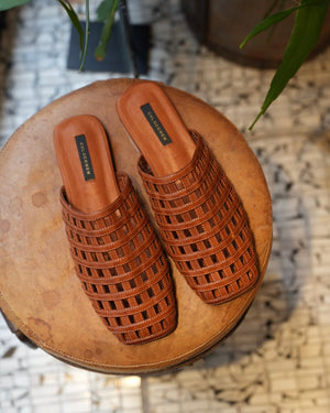 Pair of brown woven sandals on a round wooden stool with a textured floor background.
