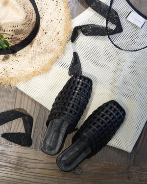 Black woven sandals on a wooden surface with a straw hat and white mesh top.