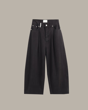 Black wide-leg cropped pants with pleated front legs, silver rivets, and a white waistband label, shown flat against a solid taupe background.