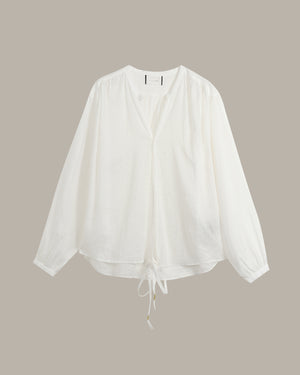 A white long-sleeve blouse with a split V-neckline and a drawstring tie at the hem, displayed against a solid taupe background.