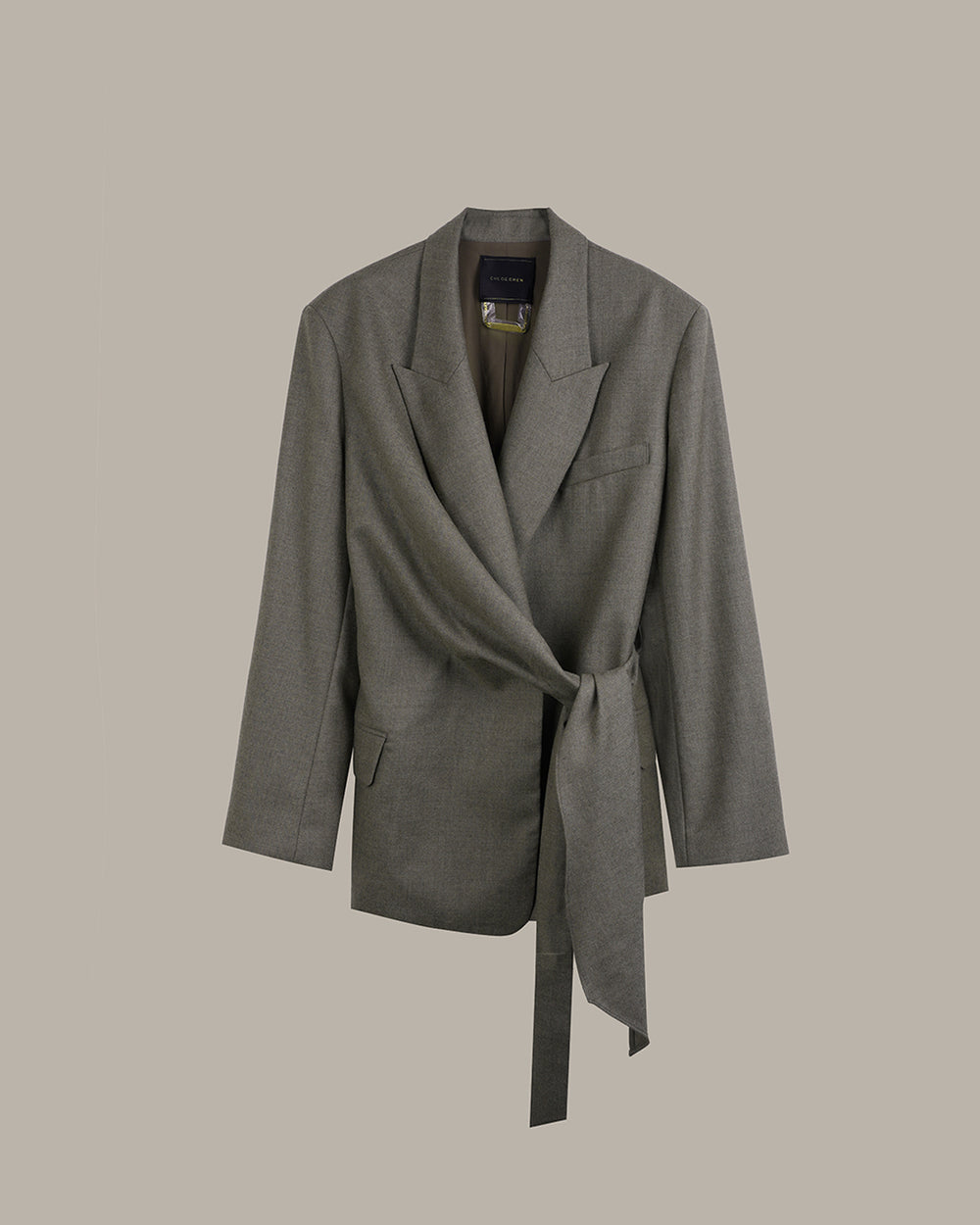 A gray wrap-style blazer with a self-tie belt, shown against a solid taupe background.