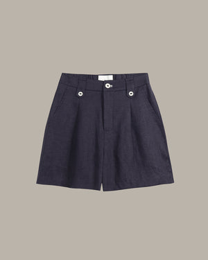 A pair of dark navy blue pleated shorts with silver button details on the front waistband, displayed flat against a solid taupe background.