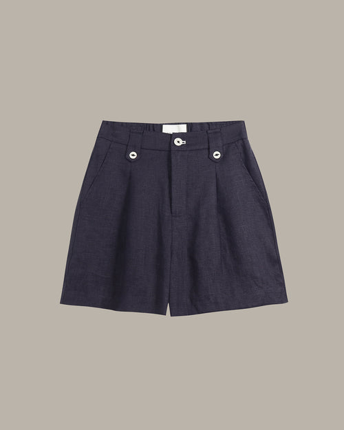 A pair of dark navy blue pleated shorts with silver button details on the front waistband, displayed flat against a solid taupe background.