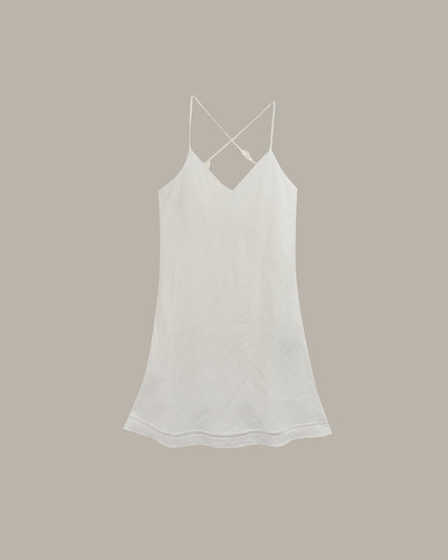 A white cotton camisole tank top with thin spaghetti straps and a crisscross back detail, shown flat against a solid taupe background.