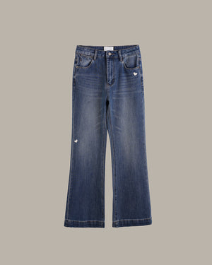 A pair of medium-wash blue denim jeans with a slight flare, featuring a small white embroidered heart on the right pocket and a small white embroidered logo on the left thigh, shown against a solid taupe background.