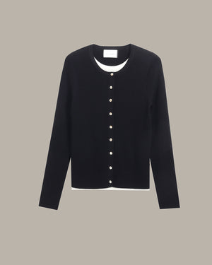 A black long-sleeve cardigan sweater with a white inner collar and a vertical row of white buttons down the front, displayed against a solid taupe background.