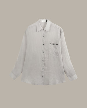 A light gray linen button-up shirt with a pointed collar, long sleeves, and a chest pocket featuring a row of small black star-shaped buttons, displayed against a solid taupe background.