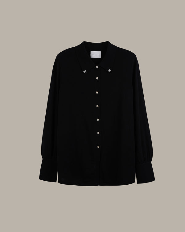 A black long-sleeve button-up shirt with small silver star embellishments on the chest and a white neck label, displayed against a solid taupe background.