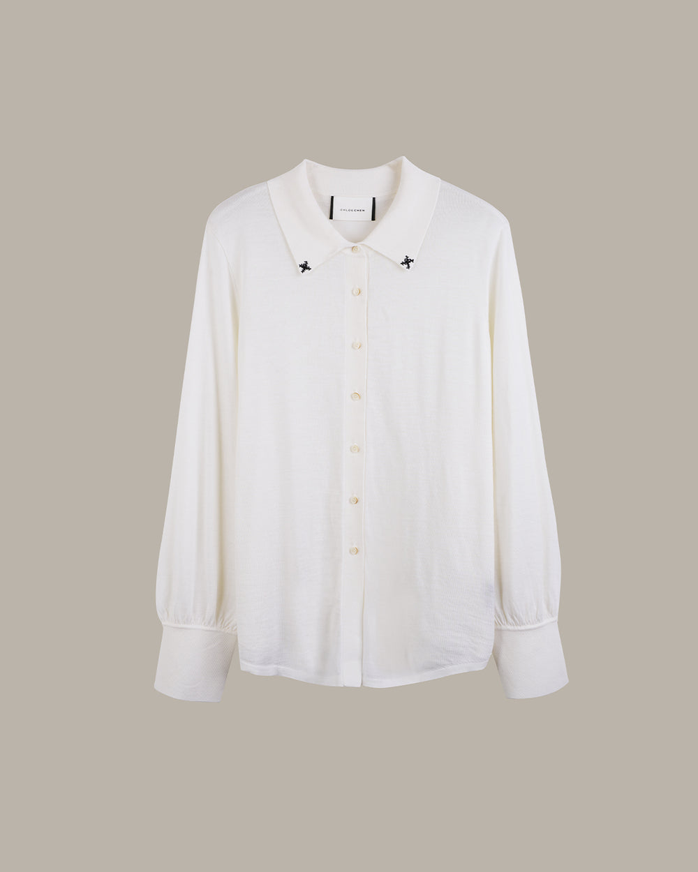 A white long-sleeve button-up shirt with a pointed collar, featuring small black embroidered star details on either side of the collar and light-colored buttons, displayed against a solid taupe background.