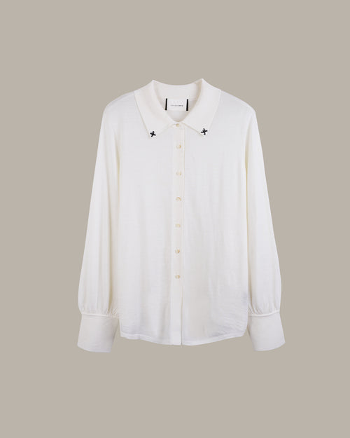 A white long-sleeve button-up shirt with a pointed collar, featuring small black embroidered star details on either side of the collar and light-colored buttons, displayed against a solid taupe background.