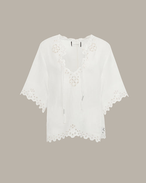 White eyelet lace short-sleeve top with a V-neckline and drawstring details, displayed against a solid taupe background.