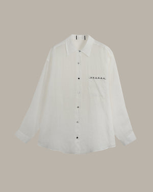 A white long-sleeve button-up shirt with a pointed collar and a small black embroidered line of stars on the left chest, displayed against a solid taupe background.