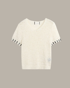 An off-white knit short-sleeve sweater with black embroidered accents along the sleeve cuffs and a small matching embroidered detail near the lower right hem, displayed against a solid taupe background.