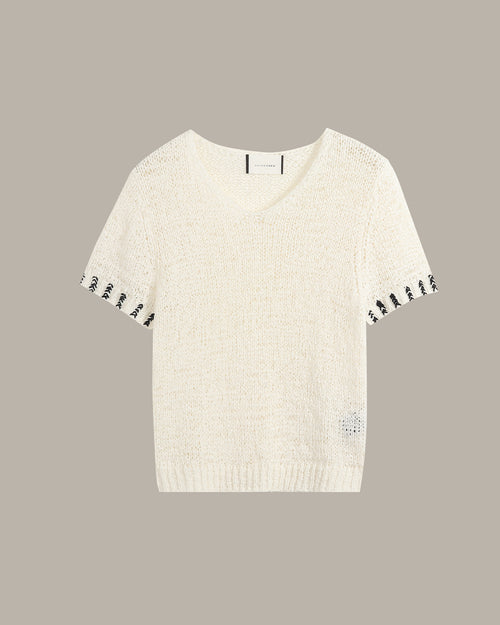 An off-white knit short-sleeve sweater with black embroidered accents along the sleeve cuffs and a small matching embroidered detail near the lower right hem, displayed against a solid taupe background.