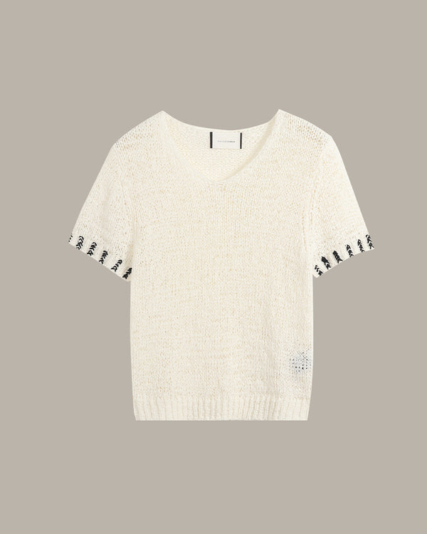 An off-white knit short-sleeve sweater with black embroidered accents along the sleeve cuffs and a small matching embroidered detail near the lower right hem, displayed against a solid taupe background.