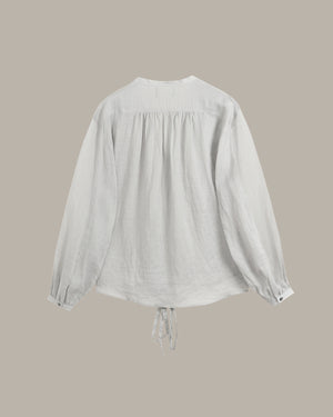 A white long-sleeve blouse with a gathered, smocked back and a drawstring tie at the hem, shown against a solid taupe background.