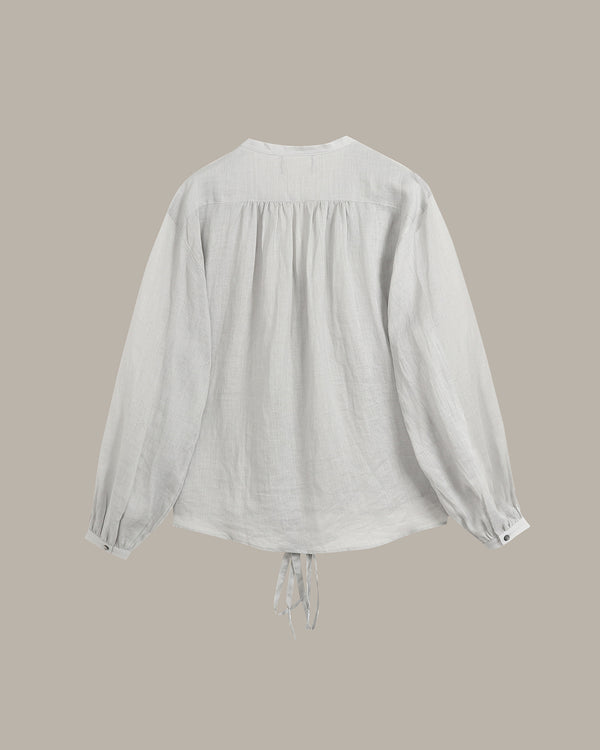 A white long-sleeve blouse with a gathered, smocked back and a drawstring tie at the hem, shown against a solid taupe background.