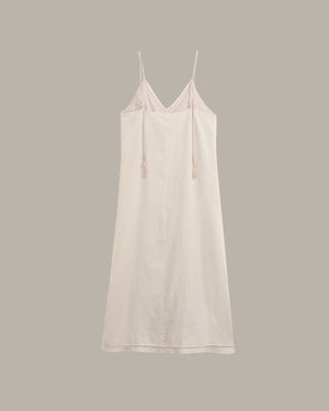 A light cream-colored cotton slip dress with thin spaghetti straps and drawstring ties, displayed against a solid taupe-gray background.