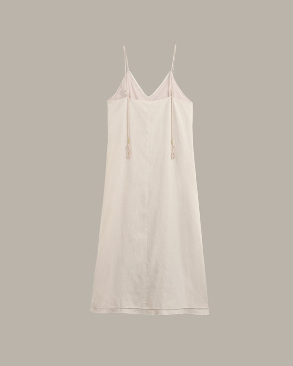 A light cream-colored cotton slip dress with thin spaghetti straps and drawstring ties, displayed against a solid taupe-gray background.