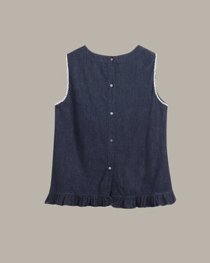 A sleeveless dark blue denim-style top with a ruffled hem, button front, and white lace trim along the armholes, shown on a plain taupe background.