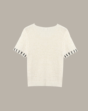 An off-white knit short-sleeve top with black-and-white patterned trim along the sleeve cuffs, shown against a solid taupe background.