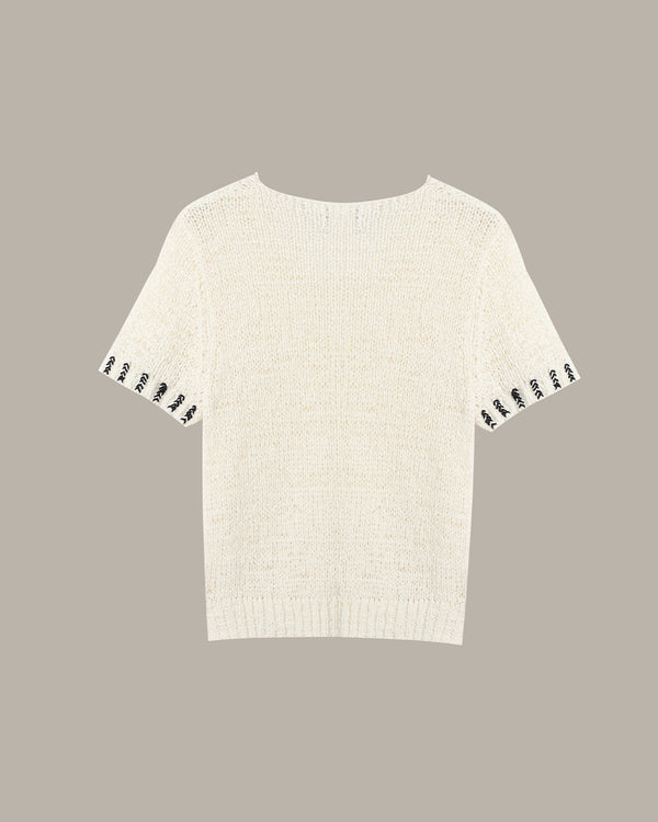 An off-white knit short-sleeve top with black-and-white patterned trim along the sleeve cuffs, shown against a solid taupe background.