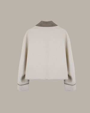The back view of a cropped, long-sleeve jacket in off-white wool, featuring a contrasting taupe suede collar and matching taupe whipstitch detailing on the cuffs, set against a solid taupe background.