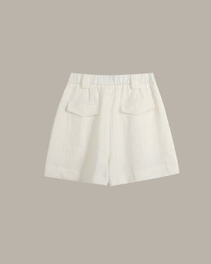 Off-white linen shorts with a pleated elastic waistband and two front patch pockets, shown flat on a solid taupe background.